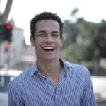 A cheerful young man in a striped shirt smiles brightly outdoors, conveying happiness and style.